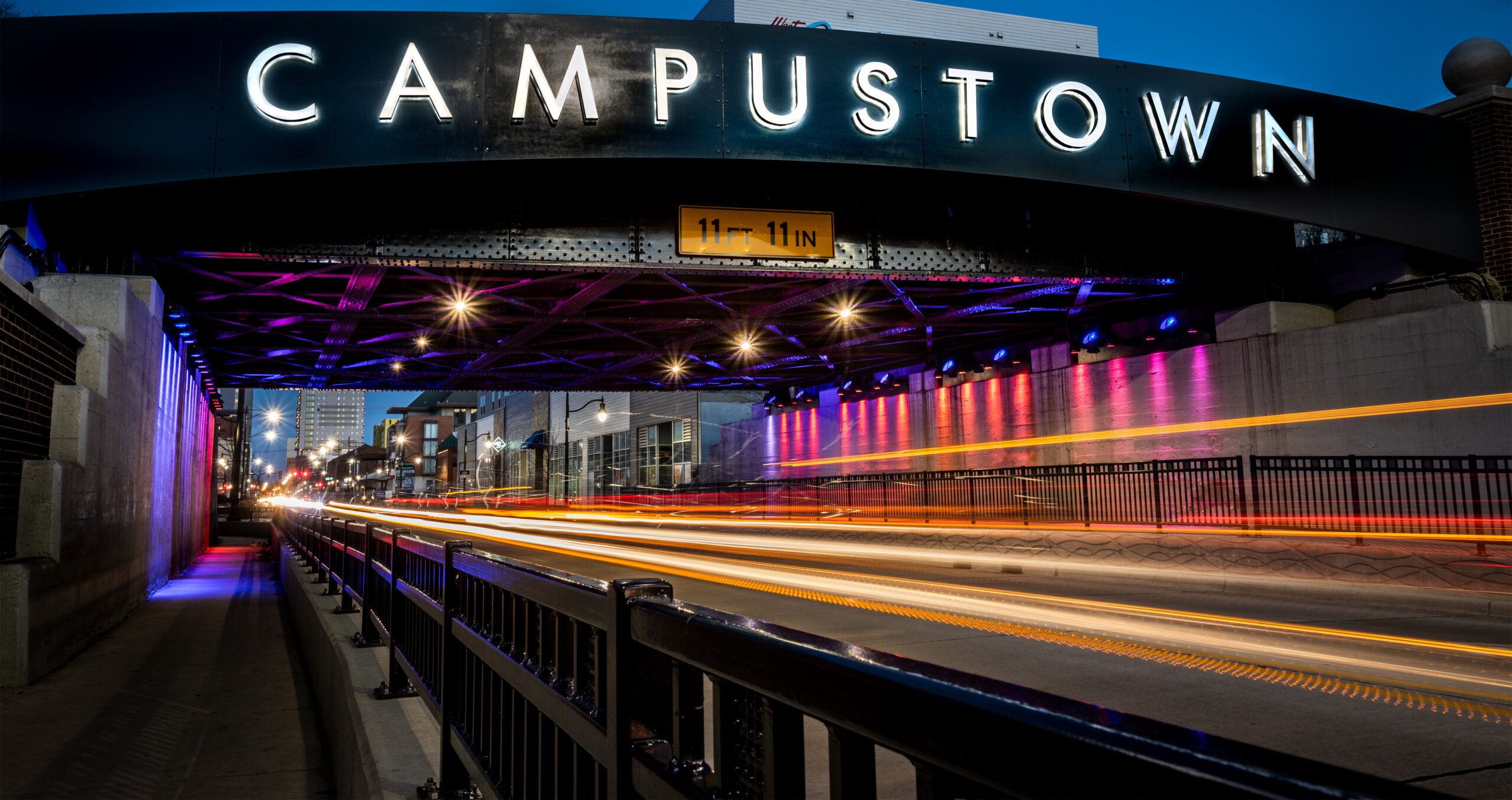 Black bridge with 'campustown' sign, neon lights, a road, and a black fence