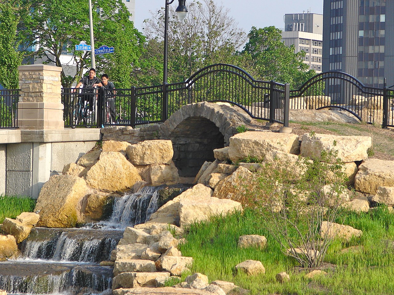 waterfall next to large rocks under a stone bridge with buildings in the background