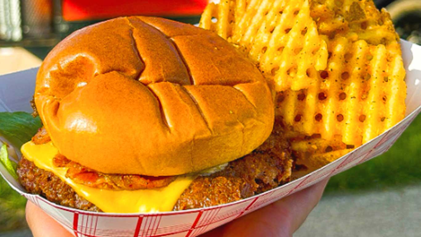 Hand holding up a paper tray of a smashburger and waffle fries