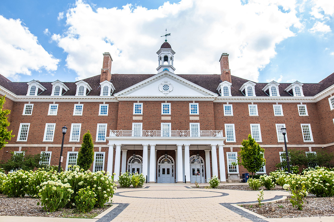 large brick building with white pillars facing a brick walkway with flowers