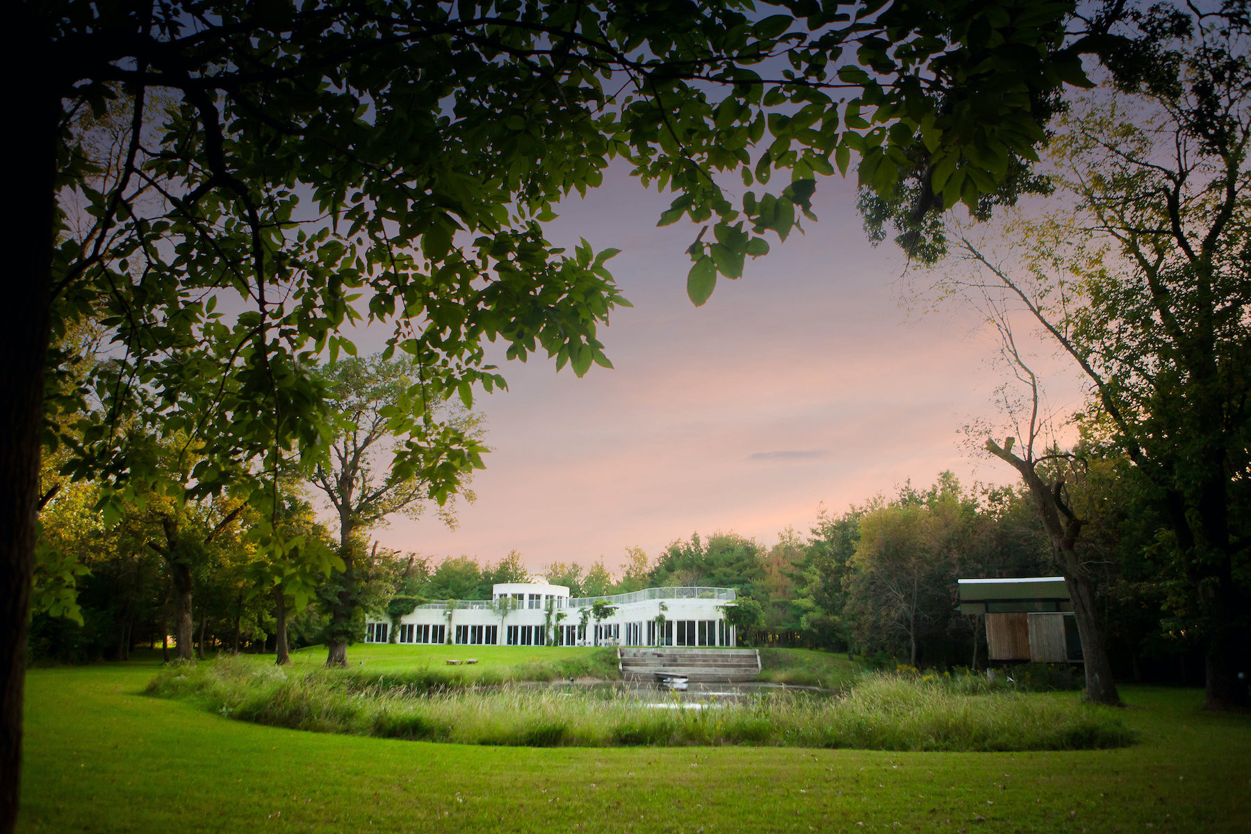 Long, white structure with many windows, surrounded by trees and a pond