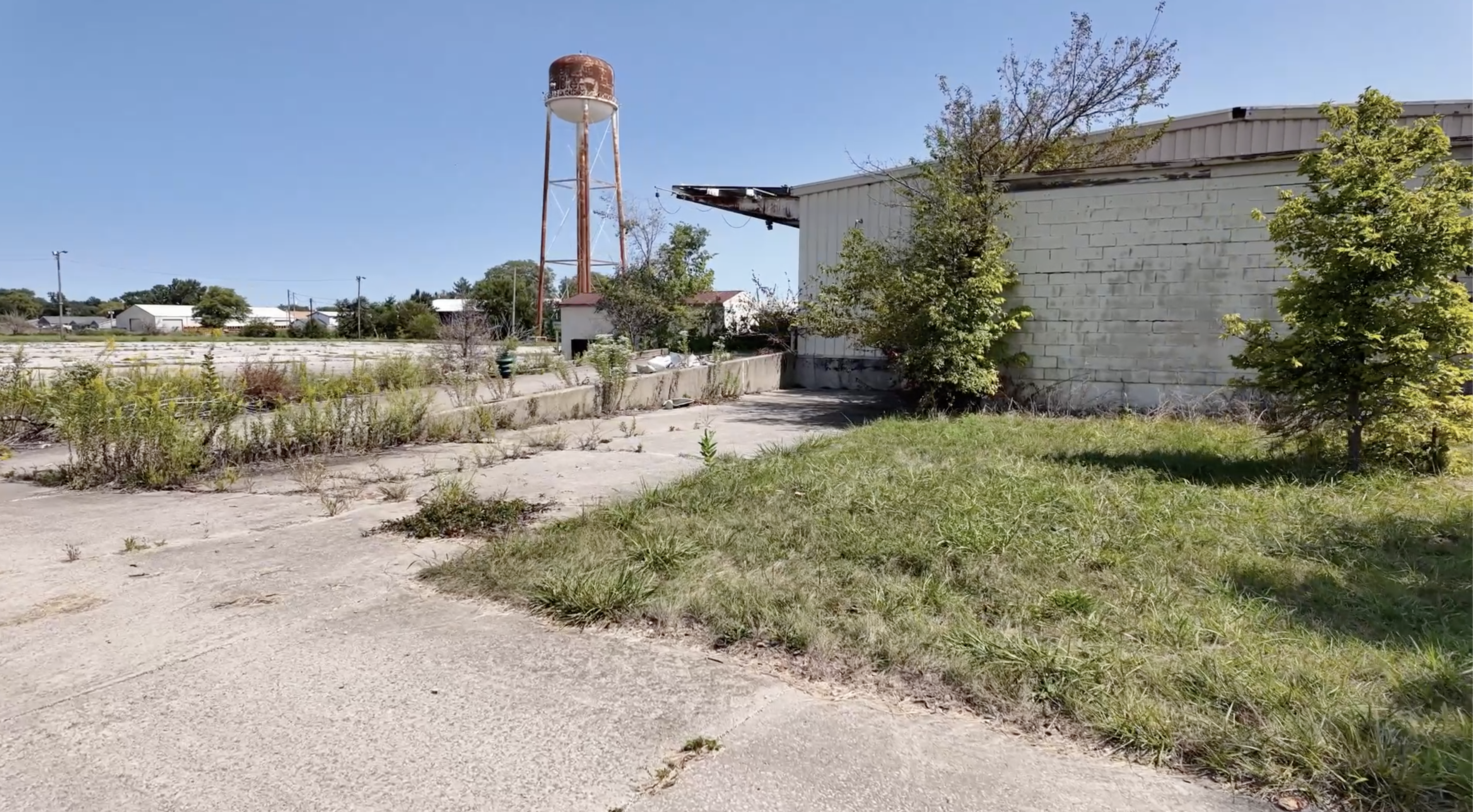 rusty water tower with overgrown parking lot and off-white brick building