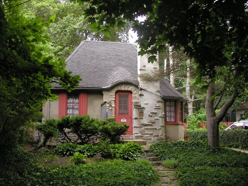 exterior of cottage with tan facade, red door and shudders, and charcoal roof, surrounded by greenery