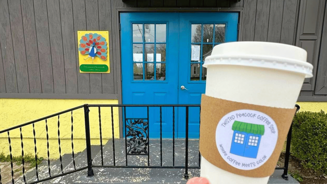 To-go cup with Twisted Peacock Coffee Shop logo held in front of the café’s bright blue double doors in Vienna, Ohio.